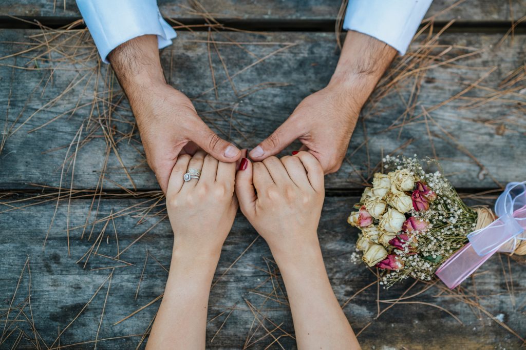 Wedding Couple Holding Hands On Wooden Table With Bouquet.